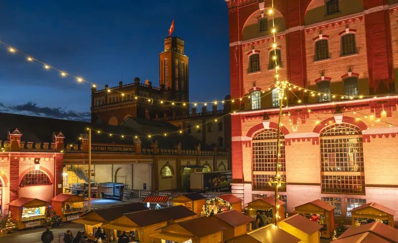 Illuminated wooden huts of the Winterdorf in front of the brick buildings and the tower of the Feldschlösschen brewery in the evening