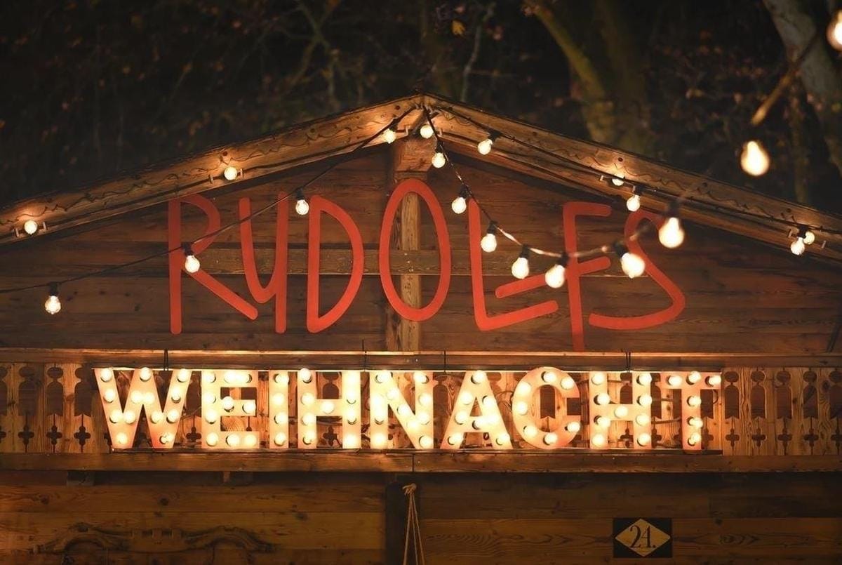 Wooden chalet with fairy lights and an illuminated «Rudolfs Weihnacht» sign in the evening